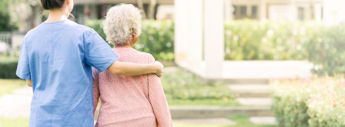 Caregiver in scrubs walking beside an older woman with a supporting arm around her.