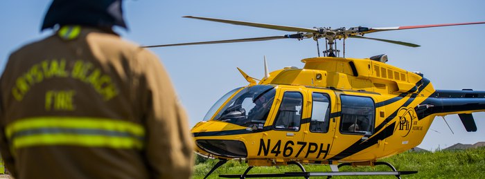 PHI Air Medical helicopter on the ground with a first responder in the foreground near a tree line.