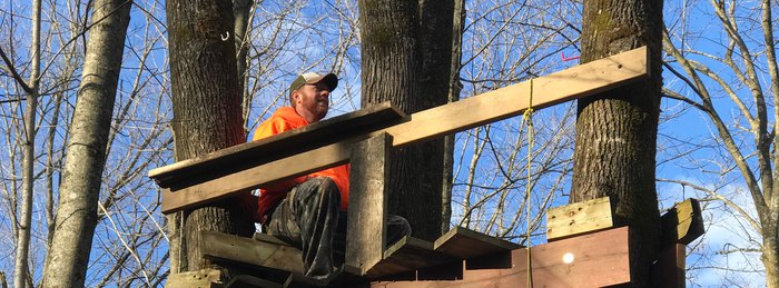 Hunter in blaze orange seated on an elevated platform stand with a haul rope hanging to the ground.
